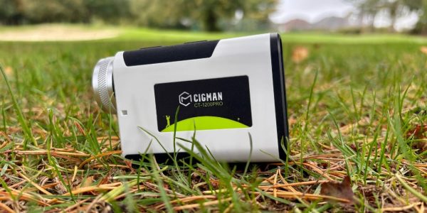 Cigman CT-1200 PRO golf laser rangefinder sitting on grass and pinestraws at golf course with bunkers and the green in the background.